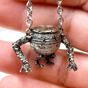 May include: A silver necklace with a distinctive pendant. The pendant is a small cauldron with arms and legs, suspended from a chain. The cauldron has detailed patterns and a textured finish, creating a whimsical design. The chain is made of small, linked rings.