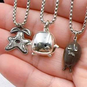 May include: Three silver-toned pendant necklaces. The pendants include a starfish, a rounded character with small legs, and a dark brown figure with small legs. The necklaces have a chain link design.