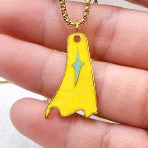 May include: A gold-colored necklace with a pendant shaped like a yellow cape. The cape pendant has a teal star design in the center. The necklace is held by a person.