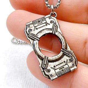 Puede incluir: Collar con colgante plateado con un diseño rectangular de estilo industrial. El colgante presenta una abertura circular central y detalles intrincados, suspendido de una cadena a juego. El collar se sostiene en una mano.