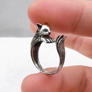May include: A silver-colored cat ring with blue eyes. The ring features a detailed cat head and paws, with the tail forming the ring's band. The cat's face is white, and the eyes are a bright blue.