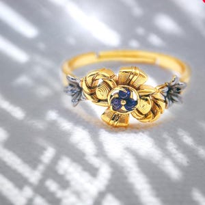 May include: A gold-toned ring featuring a floral design with a central blue stone. The flower has detailed petals and is flanked by silver-toned accents. The ring is set against a soft, light background, highlighting the jewelry's intricate details.