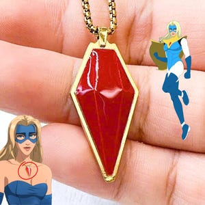 May include: A gold-colored necklace with a red, diamond-shaped pendant. The pendant has a gold border and is held by a gold chain. A cartoon image of a superheroine is in the background.