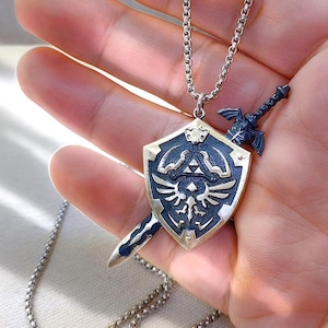 Può includere: Una collana argentata con un ciondolo raffigurante uno scudo e una spada. Lo scudo è dorato e blu navy, con un emblema dettagliato. La spada è blu navy. La collana è a catena.