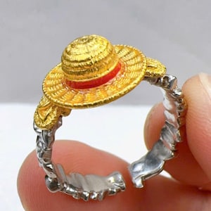 May include: A gold-coloured ring featuring a miniature straw hat design with a red band. The ring's band is silver-coloured and has a textured, open design. The ring is held between two fingers.