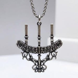 May include: A silver-toned pendant necklace featuring a three-candle menorah design. The pendant has intricate detailing, including decorative scrollwork and a textured surface. The necklace chain is a matching silver color.