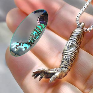 Può includere: Collana con pendente color bronzo raffigurante un braccio e una mano dettagliati. Il pendente è sospeso a una catena d'argento. Un pezzo di vetro ovale mostra un design futuristico del braccio con accenti blu e verdi.