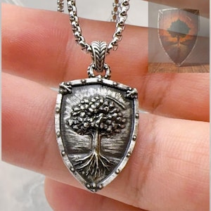 May include: A silver-toned metal pendant shaped like a shield, featuring a raised relief design of a tree with visible roots. The pendant hangs from a thick, rounded silver-toned chain. The surface has a dark, oxidised finish in the recessed areas.