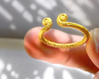 May include: A gold-colored ring with an open design. The ring features a textured surface and two curled ends. The ring is held between fingers, showcasing its intricate details and craftsmanship. The ring is a piece of jewelry.