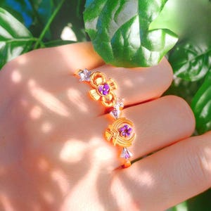 Puede incluir: Un anillo dorado con diseños florales y de media luna, acentuado con piedras preciosas moradas. El anillo presenta detalles intrincados y se encuentra sobre un fondo de follaje verde.