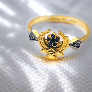May include: A gold-colored ring with a detailed design featuring a central emblem with blue gemstones. The ring has a gold band and silver accents with small purple gemstones. The ring is on a white surface.