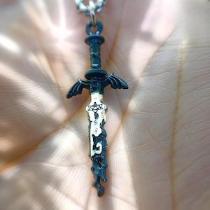 Può includere: Un ciondolo a forma di spada verde scuro con un design bianco e oro sulla lama, appeso a una catena d'argento. La spada ha un'elsa dettagliata e ali sulla guardia. Il ciondolo è lungo circa 5 cm.
