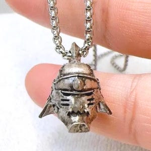 Op de afbeelding: Zilverkleurige hangerketting met een gedetailleerd helmontwerp. De hanger hangt aan een ketting. De helm heeft ingewikkelde details, waaronder gezichtskenmerken en een puntige bovenkant. De ketting wordt door een hand vastgehouden.