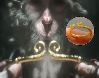 May include: A gold-colored ring with a spiral design on the ends, shown in a close-up view. The ring is held up against a blurred background of a monkey's face and a golden staff.