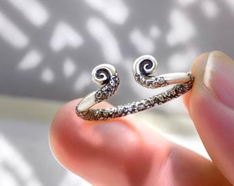 May include: A silver-colored ring with a unique design. The ring features two swirling, scroll-like ends, connected by a textured band. The ring is held between fingers, showcasing its intricate details and craftsmanship. The ring's design is reminiscent of a stylized wave or curl.