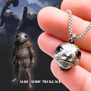 Puede incluir: Collar con colgante plateado que presenta una cabeza de criatura estilizada y detallada. El colgante cuelga de una cadena. El texto "ALBIN AURIC NECKLACE" está impreso en la parte inferior de la imagen. El fondo presenta una criatura similar.