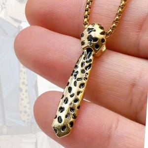 May include: Gold-toned necklace with a pendant shaped like a tie. The tie pendant features a leopard print design with black spots on a gold background. The necklace is held by a hand.