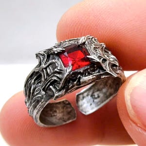 May include: A silver ring featuring a square, red gemstone. The ring has an intricate, textured design around the gem and an open band. The ring is held between fingers.
