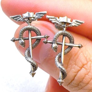 May include: A pair of silver-toned earrings featuring the Caduceus symbol. Each earring has a central staff with a serpent coiled around it, topped with wings and a star-shaped element. The earrings are held between fingers.
