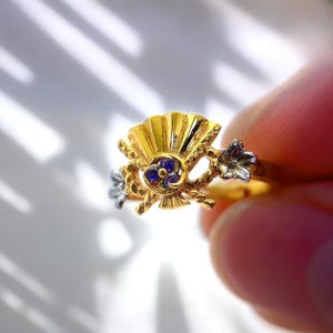 May include: A gold ring featuring a fan-shaped design with a central cluster of small blue gemstones. The ring has silver-colored floral accents on either side. The ring is held between fingers, showcasing its intricate details and craftsmanship.