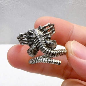 Könnte beinhalten: Silberfarbener Ring mit detailliertem, dreidimensionalem Design einer Kreatur mit skelettartigem Kopf und segmentiertem Körper. Der Ring wickelt sich mit einem spiralförmigen Band um den Finger und zeigt eine einzigartige, gotische Ästhetik.