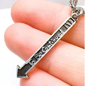Può includere: Collana con ciondolo a forma di freccia argentata con le parole "To Be Continued" incise sulla freccia. La freccia punta verso sinistra. La collana è su una catena d'argento.