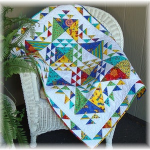 Puede incluir: Una colcha colorida con un patrón geométrico de triángulos en tonos de azul, verde, amarillo y rojo. La colcha está hecha de varias telas con diferentes patrones y texturas. Se muestra en una silla de mimbre blanca.