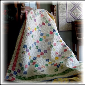 Puede incluir: Un edredón hecho a mano con un fondo blanco y un patrón de cuadrados coloridos. Los cuadrados están dispuestos en un diseño de cadena, con un borde rosa y rojo en un lado y un borde floral verde en la parte inferior. El edredón está sobre una silla de madera.