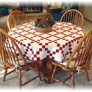 May include: A round wooden table with four chairs, all with a white and red checkered tablecloth. The table is set for a meal with a basket of greenery and pine cones in the center.