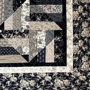 Puede incluir: Una colcha con fondo negro y un patr&oacute;n geom&eacute;trico de cuadrados de tela gris y blanco. La colcha est&aacute; bordeada con un ribete blanco y un estampado floral blanco y negro.