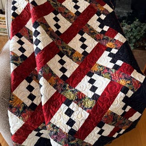 May include: A handmade quilt with a geometric pattern. The quilt features alternating red, white, and black sections, with floral fabric accents. The quilt is folded over a chair, showcasing its intricate design and color palette.