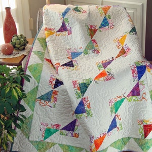 May include: A white quilt with a patchwork design featuring colorful triangles. The quilt has a green and white border with a scalloped edge. The quilt is folded and draped over a chair.