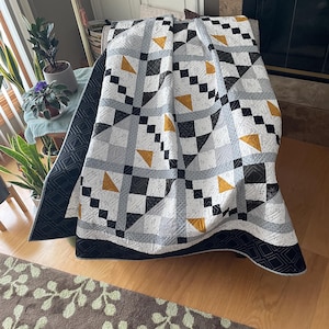 May include: A black, white, and gold quilt with a geometric pattern. The quilt is folded and draped over a couch.