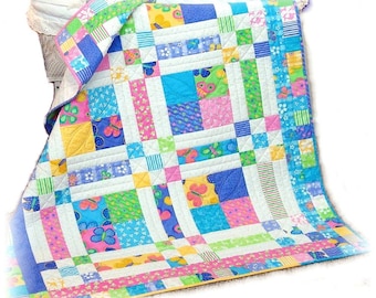 Könnte beinhalten: Eine bunte Patchwork-Decke mit einem Schmetterlingsmuster. Die Decke besteht aus verschiedenen Stoffen in Blautönen, Rosa, Gelb und Grün. Die Decke ist gefaltet und auf einer weißen Oberfläche ausgestellt.