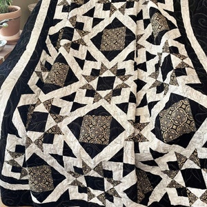 May include: A black, white, and gold quilt with a geometric pattern of stars and squares. The quilt is folded and draped over a chair.