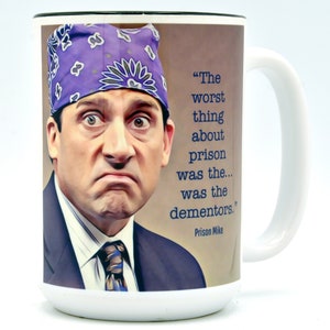 May include: White ceramic mug featuring an image of a man wearing a purple bandana, with the quote "The worst thing about prison was the... was the dementors." The mug has a white handle and rim.