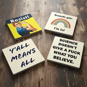 May include: Four square coasters with various text and graphics. One coaster features the text "Resist!" with a Rosie the Riveter image. Another says "Sounds gay. I'm in!" with a rainbow. The others say "Y'ALL MEANS ALL" and "SCIENCE DOESN'T GIVE A FUCK WHAT YOU BELIEVE."