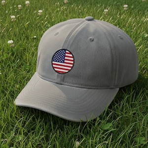 May include: A gray baseball cap with a curved brim and a circular patch featuring the American flag. The cap is resting on a bed of green grass.