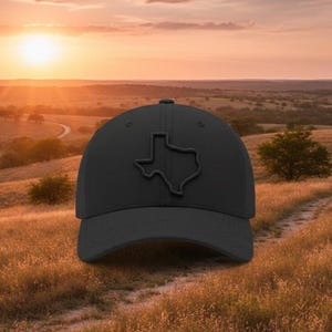 May include: A black baseball cap featuring a raised outline of the state of Texas on the front. The cap is set against a scenic background of a sunset over a grassy field.