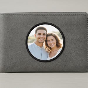 May include: Gray leather wallet with a circular photo window. The wallet features a stitched border and a black circular frame around a photo of a smiling couple. The wallet is a bi-fold style.