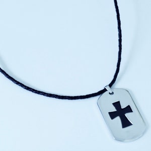 May include: A black leather cord necklace with a silver dog tag pendant featuring a black cross cutout.