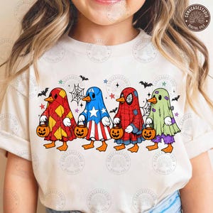 Könnte beinhalten: Ein cremefarbenes T-Shirt mit einer Grafik von vier Enten in Halloween-Kostümen. Die Enten tragen Outfits, die an einen rot-gelben Harlekin, einen blau-weißen Helden mit Sternen, ein rotes Spinnennetz-Muster und ein grünes Gespenst erinnern.