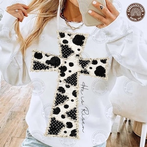 May include: White sweatshirt featuring a large cross design with a cow print pattern in black and white. The cross has a frayed edge. The words "He is Risen" are written vertically on the right side of the cross.