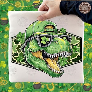 May include: A white t-shirt featuring a green dinosaur wearing a green cap and sunglasses with shamrock lenses. The dinosaur is inside a green camouflage hexagon with shamrocks around it. The design is held by a hand.