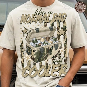 May include: A cream-colored t-shirt featuring a graphic of a camouflage-patterned cooler filled with beverage cans. The text reads "like a NORMAL DAD but COOLER" with a camouflage American flag background and several tan-colored stars.