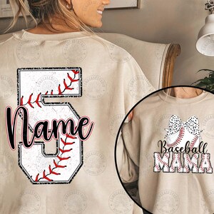 May include: A beige sweatshirt with a baseball-themed design. The back of the sweatshirt features a large number 5 with a baseball stitched into it. The front of the sweatshirt features a baseball with a bow and the text "Baseball Mama".