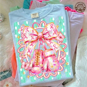 May include: A light blue t-shirt featuring a graphic of two pink boxing gloves tied together with a large pink ribbon. The design includes pink breast cancer awareness ribbons, small yellow stars, and two butterflies. The shirt has a Comfort Colors label.