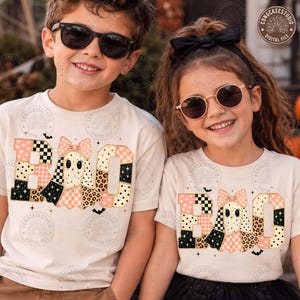 May include: Two cream-colored t-shirts featuring the word BOO in a patchwork design. The letter O is replaced by a ghost wearing a bow. The letters feature patterns including black and white checkers, leopard print, polka dots, and stars.