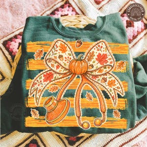 May include: A dark green sweatshirt featuring a graphic of a stethoscope with an orange pumpkin at the center of the chest piece. The stethoscope is adorned with a large bow patterned with autumn leaves and acorns over orange horizontal stripes.