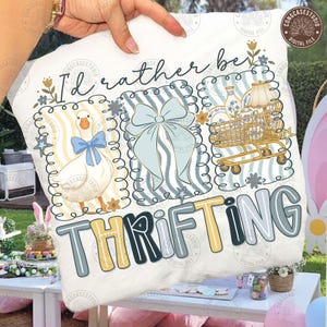 Puede incluir: Un cojín blanco con la frase "I'd rather be thrifting" en una fuente lúdica. El diseño incluye ilustraciones de un ganso, un lazo y un carrito de compras, todo en colores pastel suaves. El cojín es sostenido por una persona.